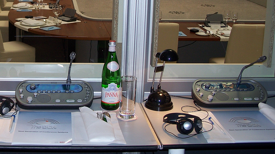 Beschreibung: Ein Blick durch eine Dolmetscherkabine zeigt im Vordergrund zwei Dolmetscherarbeitsplätze und im Hintergrund eine für ein Abendessen vorbereitete Veranstaltung für Führungskräfte (Executive Dinner).